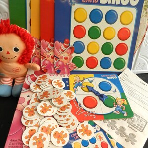 Pode incluir: Um conjunto de jogo de Bingo Candy Land vintage. O jogo inclui um tabuleiro de bingo azul com círculos coloridos, fichas de homem de gengibre e cartões de instruções. Uma boneca Raggedy Ann é visível no lado esquerdo da imagem.