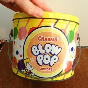 Puede incluir: Una lata de metal amarilla con asa, con un diseño colorido de piruletas y el texto "CHARMS Blow Pop Juniors".