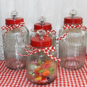 May include: Four clear glass jars with red lids and white and red gingham ribbon tied around the jars. The jars have a crystal knob on the lid. One jar is filled with colorful candies.