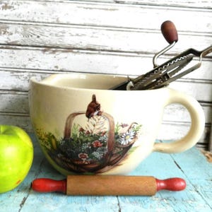 以下が含まれることがあります： クリーム色のセラミック製ミキシングボウル。花柄のバスケットのデザイン。ヴィンテージの泡立て器と、赤いハンドルが付いた小さな木製のめん棒がボウルの中と前にあります。左側には青リンゴがあります。