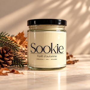 Peut inclure: Une bougie en pot de verre transparent avec un couvercle noir et une étiquette crème portant le texte Sookie, Forêt d'automne et 270 ml. La bougie contient de la cire blanc cassé et est placée devant des aiguilles de pin et une pomme de pin.