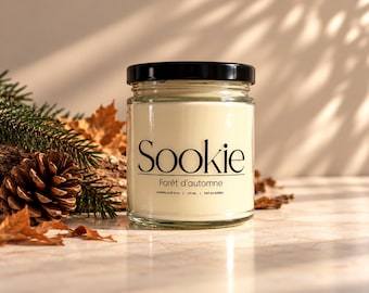 May include: A clear glass jar candle with a black lid and a cream-coloured label featuring the text Sookie, Forêt d'automne, and 270 ml. The candle contains off-white wax and is positioned in front of pine needles, a pinecone, and dried leaves.