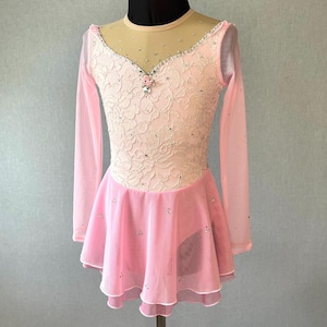 May include: A light pink figure skating dress with long sleeves. The bodice features a cream-colored lace overlay with rhinestone and floral accents. The skirt is a layered pink chiffon with scattered rhinestones. The dress is displayed on a mannequin.