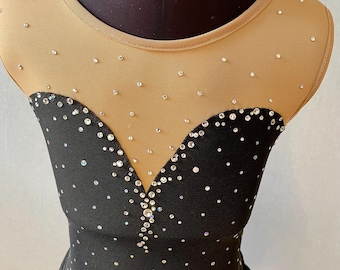 May include: Close-up of a black leotard with a nude mesh neckline and upper chest. The leotard is embellished with scattered rhinestones and a heart-shaped design of rhinestones at the bodice. The garment is likely for dance or figure skating.