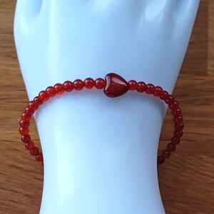 Peut inclure: Bracelet de perles rouges avec un charm en forme de cœur. Le bracelet est composé de petites perles rondes rouges et comporte une perle plus grande en forme de cœur au centre. Le bracelet est présenté sur une surface blanche.