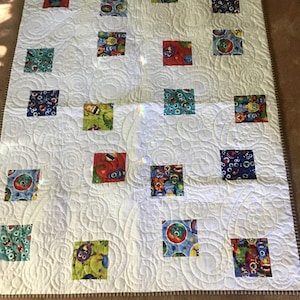 May include: A white quilt with a repeating pattern of colorful square patches. Each square features a different whimsical monster design. The quilt has a stitched spiral pattern and a brown border.