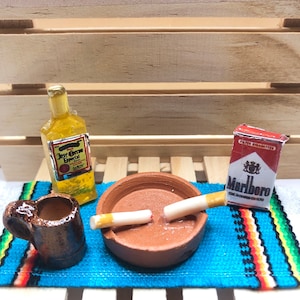 May include: A miniature scene with a brown ceramic mug, a clear glass bottle with a label that reads "Jose Cuervo Especial", a terracotta ashtray with two cigarettes, and a pack of Marlboro cigarettes on a blue and red striped rug.