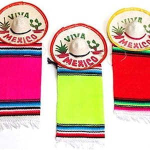May include: Three miniature sombreros with matching serape scarves. Each sombrero has the words "VIVA MEXICO" and a cactus design. The serapes are bright pink, green, and red with colorful stripes and fringe.