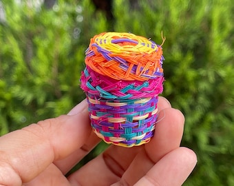 Könnte beinhalten: Ein kleiner, bunter geflochtener Korb aus leuchtend bunten Plastikfäden. Der Korb ist in einem engen Muster geflochten und hat eine zylindrische Form. Die Farben des Korbs sind Gelb, Orange, Rosa, Lila und Grün.