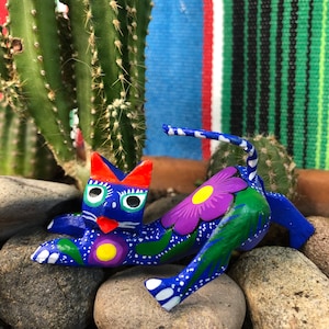 K&ouml;nnte beinhalten: Eine handbemalte Holzkatzenfigur mit blauen, gr&uuml;nen, wei&szlig;en, gelben, violetten und orangefarbenen Details. Die Katze liegt auf der Seite mit eingerolltem Schwanz. Die Figur ist mit Blumen und Bl&auml;ttern verziert.