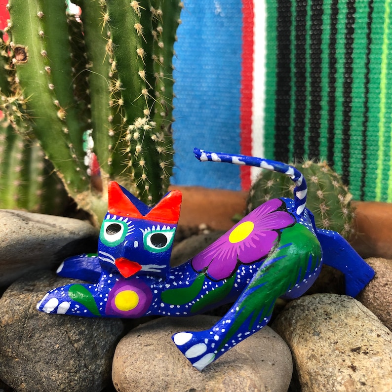 K&ouml;nnte beinhalten: Eine handbemalte Holzkatzenfigur mit blauen, gr&uuml;nen, wei&szlig;en, gelben, violetten und orangefarbenen Details. Die Katze liegt auf der Seite mit eingerolltem Schwanz. Die Figur ist mit Blumen und Bl&auml;ttern verziert.