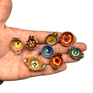 May include: Eight miniature ceramic bowls with painted floral designs. The bowls are brown, red, blue, and yellow with white accents. The bowls have a small handle on the side.