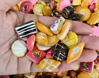 Könnte beinhalten: Eine Hand hält eine Auswahl an bunten und gemusterten Mini-Backwaren, darunter Kekse, Kuchen und Gebäck. Die Backwaren sind in einem zufälligen Muster angeordnet und haben alle unterschiedliche Formen und Größen.