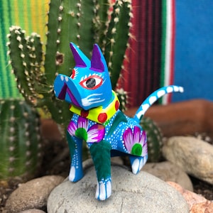 K&ouml;nnte beinhalten: Eine blaue Holzkatzenfigur mit wei&szlig;en, gelben, gr&uuml;nen und violetten Maldetails. Die Katze steht auf einem Stein mit einem Kaktus im Hintergrund.
