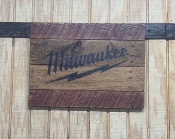 May include: A wooden sign with the word "Milwaukee" in black lettering and a lightning bolt design. The sign is made of reclaimed wood and has a rustic look.
