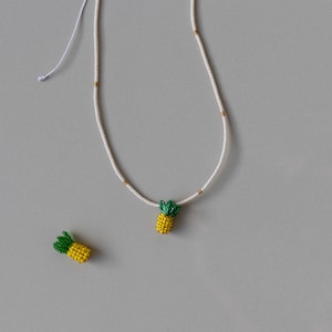 May include: A white beaded necklace with a yellow and green pineapple pendant. A matching pineapple charm is next to the necklace. The necklace has gold-colored accents and a white string closure. The background is a solid gray.