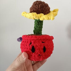 Pode incluir: Um girassol de crochê feito à mão em um vaso vermelho. O girassol tem um centro marrom, pétalas amarelas e um caule verde. O vaso tem um rosto sorridente com olhos pretos e uma boca pequena.