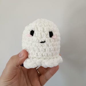 May include: A small, white, crocheted ghost with black eyes and a small mouth. The ghost has pink blush on its cheeks and a ruffled bottom edge. The plush toy is handmade and has a soft, textured appearance.