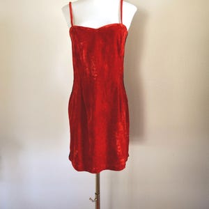May include: A vibrant red velvet dress with thin straps and a sweetheart neckline. The dress is displayed on a white mannequin, showcasing its form-fitting silhouette. The dress is approximately 86 cm long.