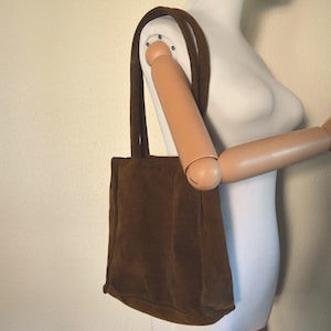 May include: A brown suede tote bag with long straps, displayed on a mannequin. The bag is rectangular with a simple design, and the straps are attached at the top. The bag is worn over the shoulder.
