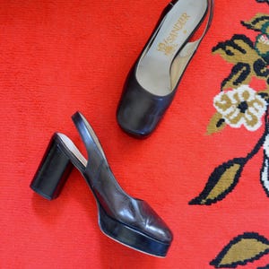 Könnte beinhalten: Ein Paar dunkelbraune Slingback-Schuhe aus Leder mit quadratischer Zehenpartie, dicker Plateausohle und klobigem Blockabsatz. Die Innensohle zeigt den Markennamen SANDLER in goldener Schrift auf einem hellen Hintergrund.
