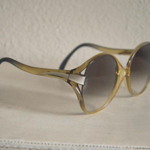 May include: Vintage oversized sunglasses with a light brown frame and gradient lenses. The arms are black, and the frame has a silver accent. These sunglasses offer a retro aesthetic.