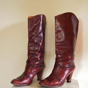 Könnte beinhalten: Ein Paar hohe, weinrote Lederstiefel mit einem herzförmigen Design in der Nähe des oberen Teils. Die Stiefel haben eine spitze Zehenpartie und einen hohen Absatz. Die Stiefel werden auf einer weißen Oberfläche präsentiert.