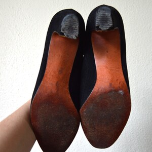 May include: A pair of black suede heeled shoes with brown leather soles. The shoes are shown from the bottom, with the soles facing the camera.