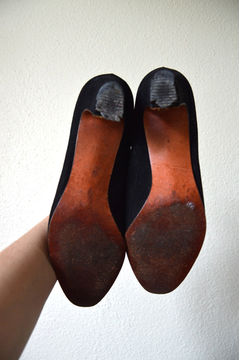 May include: A pair of black suede heeled shoes with brown leather soles. The shoes are shown from the bottom, with the soles facing the camera.