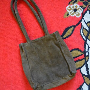 May include: A brown suede tote bag with two matching handles. The bag is rectangular with a slightly rounded bottom. The bag is set against a red background with a floral pattern in the corner.