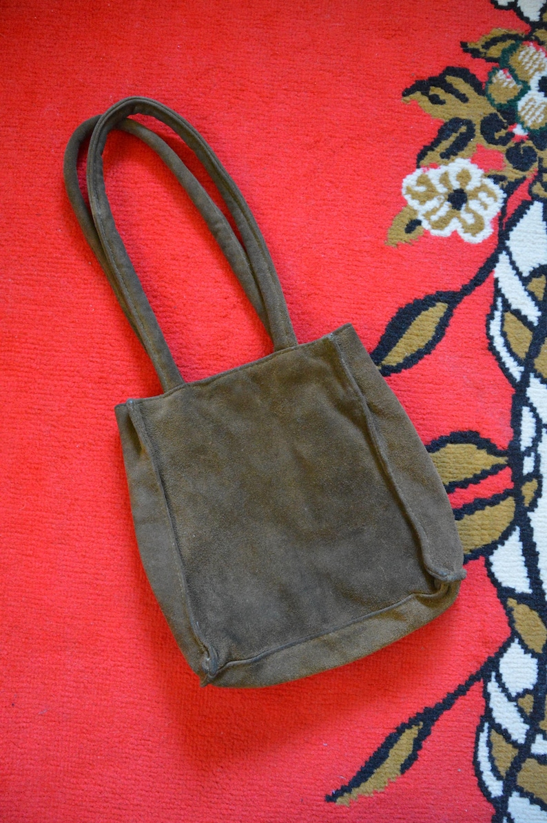May include: A brown suede tote bag with two matching handles. The bag is rectangular with a slightly rounded bottom. The bag is set against a red background with a floral pattern in the corner.