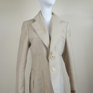 Puede incluir: Una chaqueta beige con cierre de un solo botón y tres botones dorados. La chaqueta tiene un bolsillo de parche en el lado izquierdo y un bolsillo con solapa en el lado derecho.