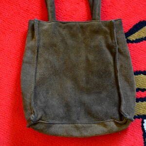 May include: A brown suede tote bag with two handles. The bag is rectangular with a slightly rounded bottom and is set against a red background.
