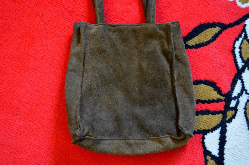 May include: A brown suede tote bag with two handles. The bag is rectangular with a slightly rounded bottom and is set against a red background.