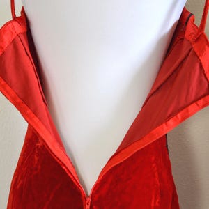 May include: Close-up of a red velvet dress with a zip closure. The dress has thin straps and a satin lining. The zip is partially open, revealing the interior lining and construction of the garment.