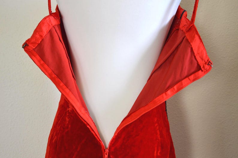 May include: Close-up of a red velvet dress with a zip closure. The dress has thin straps and a satin lining. The zip is partially open, revealing the interior lining and construction of the garment.