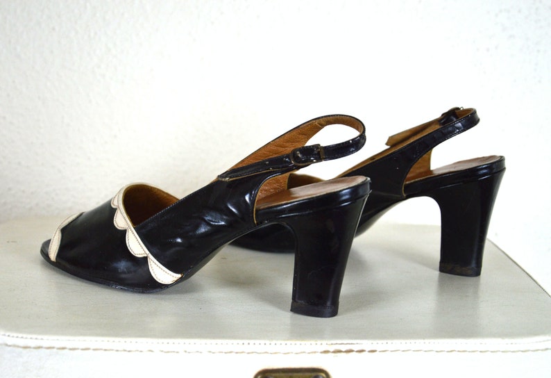 May include: A pair of black heeled shoes with white scalloped trim around the toe. The shoes have a slingback strap with a buckle closure.