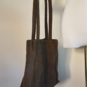 May include: A brown suede tote bag with four long straps. The bag is held up by a mannequin arm. The bag has a rectangular shape with a rounded bottom. The suede material has a textured appearance.