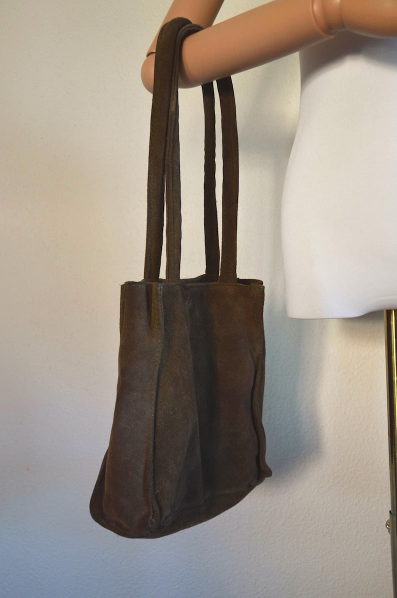 May include: A brown suede tote bag with four long straps. The bag is held up by a mannequin arm. The bag has a rectangular shape with a rounded bottom. The suede material has a textured appearance.