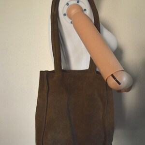 May include: A brown suede tote bag with two shoulder straps. The bag is rectangular with a simple design and a soft, textured appearance. The bag is displayed on a mannequin.