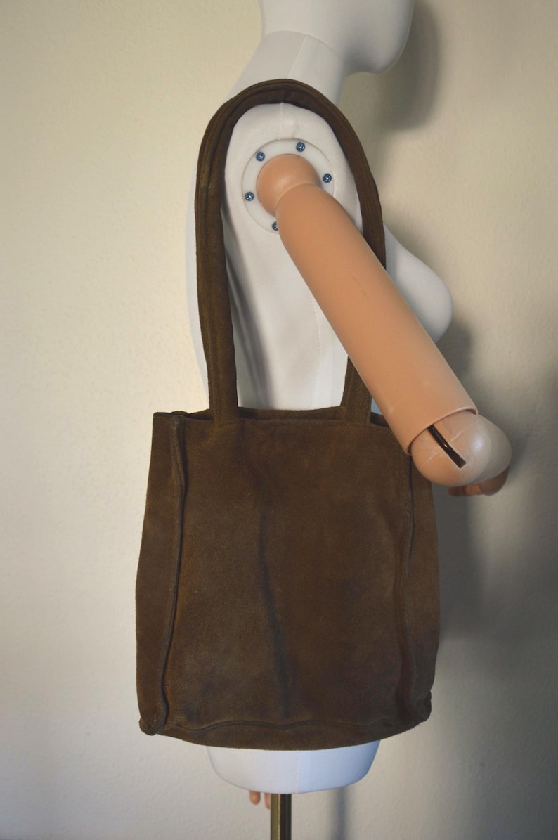 May include: A brown suede tote bag with two shoulder straps. The bag is rectangular with a simple design and a soft, textured appearance. The bag is displayed on a mannequin.