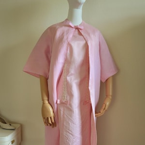 May include: A light pink vintage dress and jacket set displayed on a mannequin. The dress is sleeveless with a decorative lace panel and bow accents. The matching jacket has short sleeves and a bow at the neckline. The outfit is made of a silky fabric.