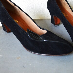 May include: A pair of black suede heeled shoes with a small black leather accent on the toe. The shoes have a brown heel and are on a gray surface.