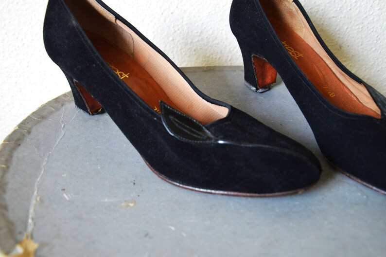 May include: A pair of black suede heeled shoes with a small black leather accent on the toe. The shoes have a brown heel and are on a gray surface.