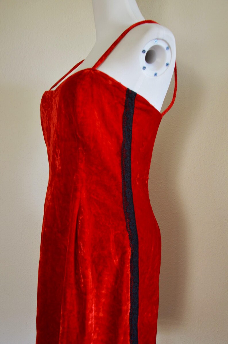 May include: A red velvet dress with spaghetti straps and a dark blue lace trim down the side. The dress is displayed on a white mannequin. The fabric has a textured appearance.