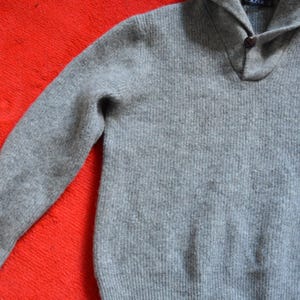 Puede incluir: Un suéter gris con cuello chal y cierre de un solo botón. El suéter tiene mangas largas y una textura acanalada. La palabra "BLAZER" es visible en la etiqueta. El suéter se muestra sobre una superficie roja.
