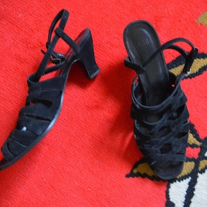 May include: A pair of black suede sandals with a strappy design and small heels. The sandals have ankle straps and are set against a red background.
