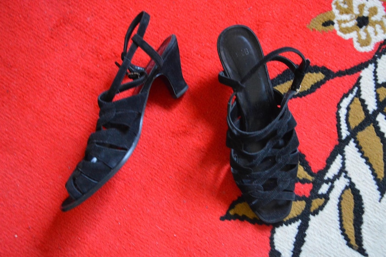 May include: A pair of black suede sandals with a strappy design and small heels. The sandals have ankle straps and are set against a red background.