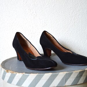 May include: A pair of black suede heeled shoes with a pointed toe and a decorative detail on the vamp. The shoes are sitting on a striped box.