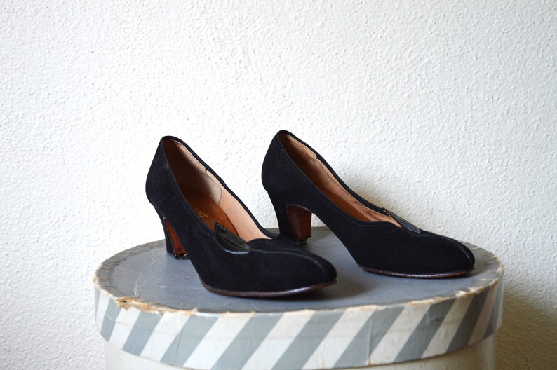 May include: A pair of black suede heeled shoes with a pointed toe and a decorative detail on the vamp. The shoes are sitting on a striped box.